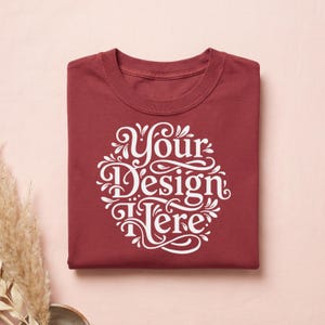 May include: A folded maroon t-shirt with a white, ornate design that reads "Your Design Here." The shirt is made of a soft material and is neatly folded. The background is a light pink color.