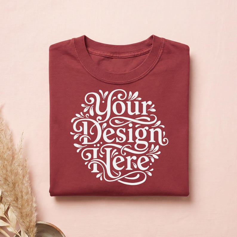 May include: A folded maroon t-shirt with a white, ornate design that reads "Your Design Here." The shirt is made of a soft material and is neatly folded. The background is a light pink color.