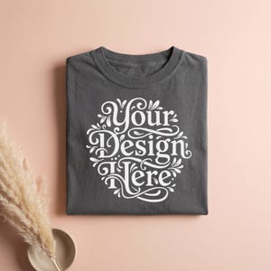 May include: A folded charcoal gray t-shirt with the text "Your Design Here" in a white, ornate, circular design. The shirt is centered on a light pink surface, with a decorative plant and a small bowl in the lower left corner.