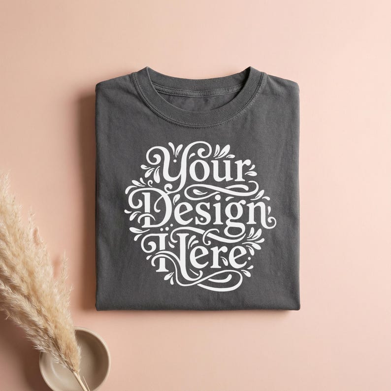 May include: A folded charcoal gray t-shirt with the text "Your Design Here" in a white, ornate, circular design. The shirt is centered on a light pink surface, with a decorative plant and a small bowl in the lower left corner.