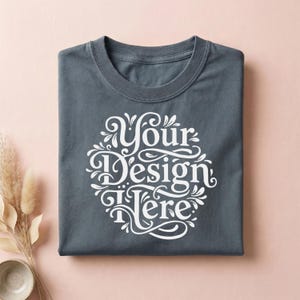 May include: A folded, slate-gray t-shirt with a white, ornate design that reads "Your Design Here." The design is circular and features decorative flourishes. The shirt is on a pink surface with dried plants and a small bowl.