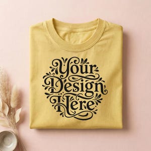 May include: A folded, mustard-yellow t-shirt with a crew neck. The shirt features a black, ornate design that reads "Your Design Here" in a decorative font. The shirt is neatly folded on a pink surface.