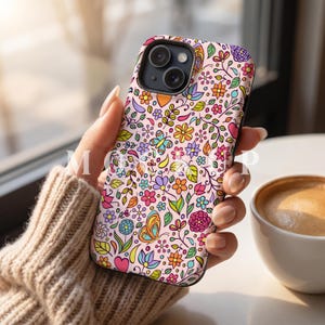 May include: A phone case with a floral design, showcasing a pattern of flowers, butterflies, and leaves in various colours on a pale pink backdrop. The case has a black edge. A cup of coffee is in the background.
