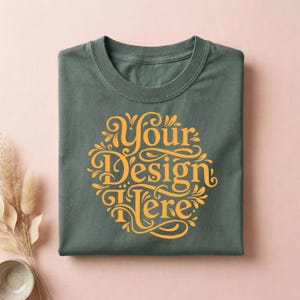 May include: A folded, dark green t-shirt with a round, ornate, yellow design that reads "Your Design Here". The shirt is made of a soft material and is displayed on a pink surface.