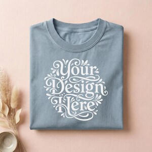 May include: A folded, light blue t-shirt with a white, ornate design that reads "Your Design Here." The shirt is made of a soft material and has a crew neck. The shirt is displayed on a pink surface.