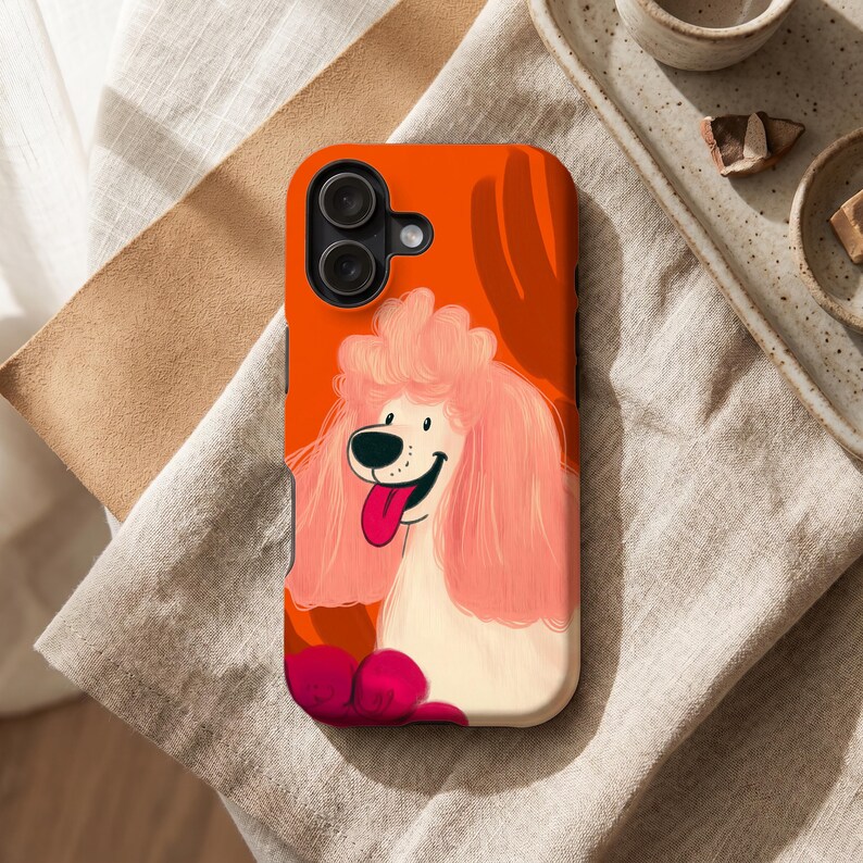 Peut inclure: Coque de t&eacute;l&eacute;phone avec un dessin de caniche rose et une langue rouge sur fond orange. Le bo&icirc;tier comporte un module cam&eacute;ra noir et repose sur une surface beige.