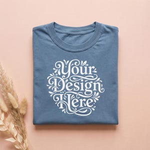 May include: A folded, dusty blue t-shirt with the text "Your Design Here" in a white, ornate, circular design. The shirt is centered on a light pink surface, with dried ornamental grass on the left.