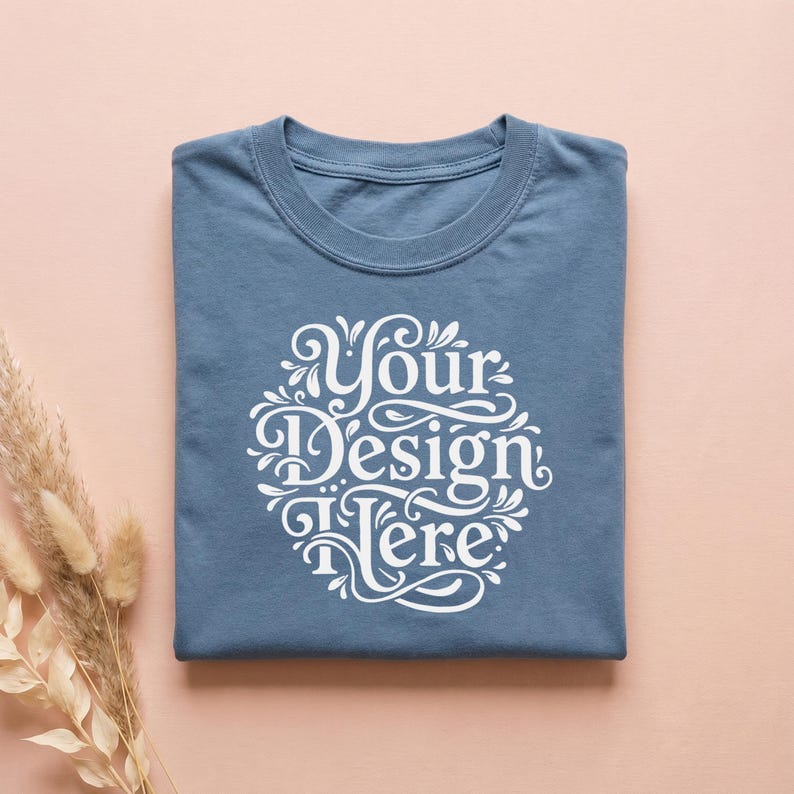 May include: A folded, dusty blue t-shirt with the text "Your Design Here" in a white, ornate, circular design. The shirt is centered on a light pink surface, with dried ornamental grass on the left.
