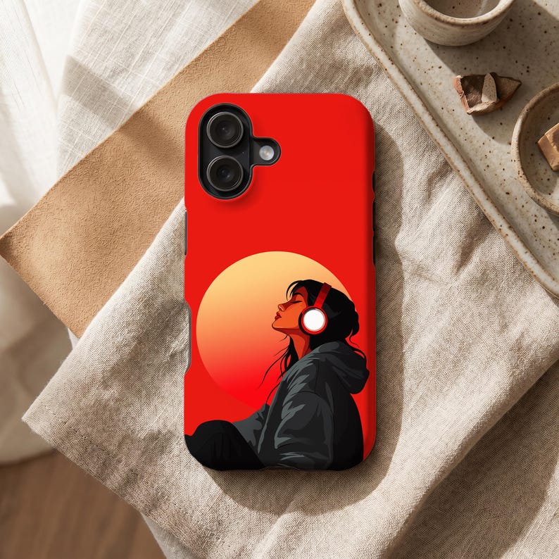 Peut inclure: Coque de t&eacute;l&eacute;phone rouge avec un graphisme d'une personne portant un casque, sur fond de coucher de soleil. La personne porte un sweat &agrave; capuche noir. La coque a un appareil photo &agrave; double objectif.