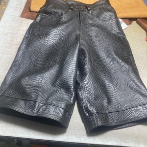 May include: Black leather shorts featuring a textured snakeskin pattern, a high-waisted design with a button closure, front pockets, and wide legs with a stitched hem detail. The material has a glossy finish and a distinct reptilian scale texture.