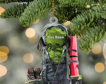 May include: A Christmas ornament featuring a green and gray backpack with the text "Your Name", hiking boots, trekking poles, a red sleeping pad, and a camera. The ornament hangs from a gold chain.