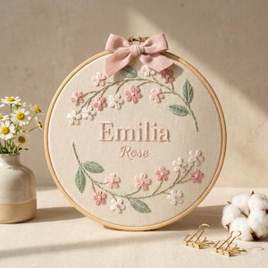 May include: A round, cream-colored embroidery hoop featuring the name "Emilia Rose" in pink thread, surrounded by a floral wreath of pink and white flowers and green leaves. A pink bow adorns the top. The hoop is set on a light surface.