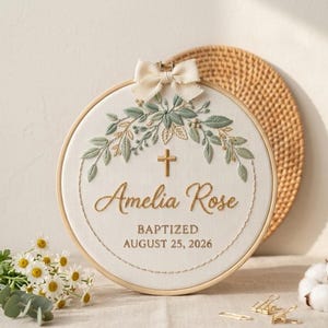 May include: Embroidered baptism hoop art featuring the name "Amelia Rose", a cross, and the date "August 25, 2026". The design includes a floral wreath in green and beige, with a cream-colored bow at the top.