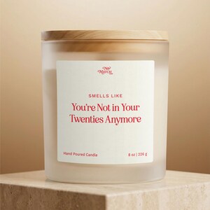 Peut inclure: Une bougie en verre dépoli avec un couvercle en bois clair. L'étiquette blanche porte le texte rouge No Match, SMELLS LIKE, You're Not in Your Twenties Anymore, Hand Poured Candle et 8 oz | 226 g. La cire est visible à l'intérieur du pot.