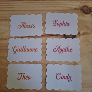 May include: Six white place cards with scalloped edges, each bearing a name in a cursive font. Names include Alexis, Sophia, Guillaume, Agathe, Théo, and Cindy. The names are in shades of orange and pink.