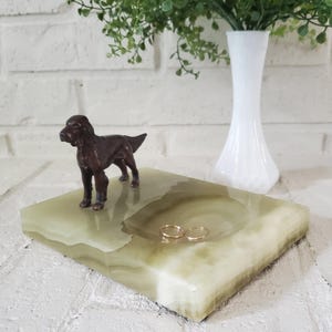 May include: A rectangular light green stone tray featuring a small brown dog figurine standing on one side and a circular indentation in the center. Two thin gold-colored metal rings are placed inside the circular depression of the stone tray.