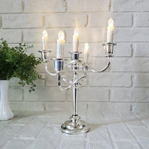 May include: A silver-toned metal candelabra featuring five arms with white electric taper candles. The central stem is elevated, surrounded by curved arms of varying heights, all supported by a circular base. The candles emit a soft glow.
