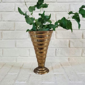 May include: A vintage brass vase with a fluted design, holding fresh green foliage. The vase has a wide top and a narrow, tiered base. The brass has a warm, aged patina, adding to its antique charm. The vase is a decorative item.