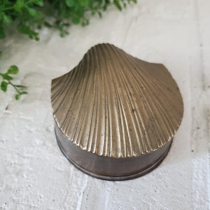 May include: A brass trinket box shaped like a scallop shell. The lid features a textured, radiating ribbed pattern that mimics the natural ridges of a seashell. The base is a smooth, circular metal band that supports the shell-shaped top cover.
