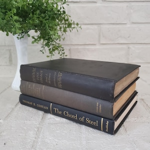 May include: A stack of three hardcover books with dark covers. The top book is black, the middle is grey, and the bottom book is black with gold text reading The Chord of Steel by Thomas B. Costain and the publisher name Doubleday.