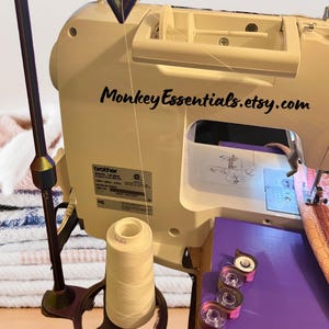 May include: A sewing machine with a white thread spool and a thread stand with a metal arm. The thread stand has a wheel to hold the thread spool. There are three spools of thread on the table. The sewing machine is a Brother model LB-6800.