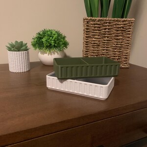 May include: Two decorative rectangular containers, one white and one olive green, are stacked on a dark wood surface. The white container has vertical ribbed detailing. The green container has two internal compartments. Artificial plants in pots are in the background.