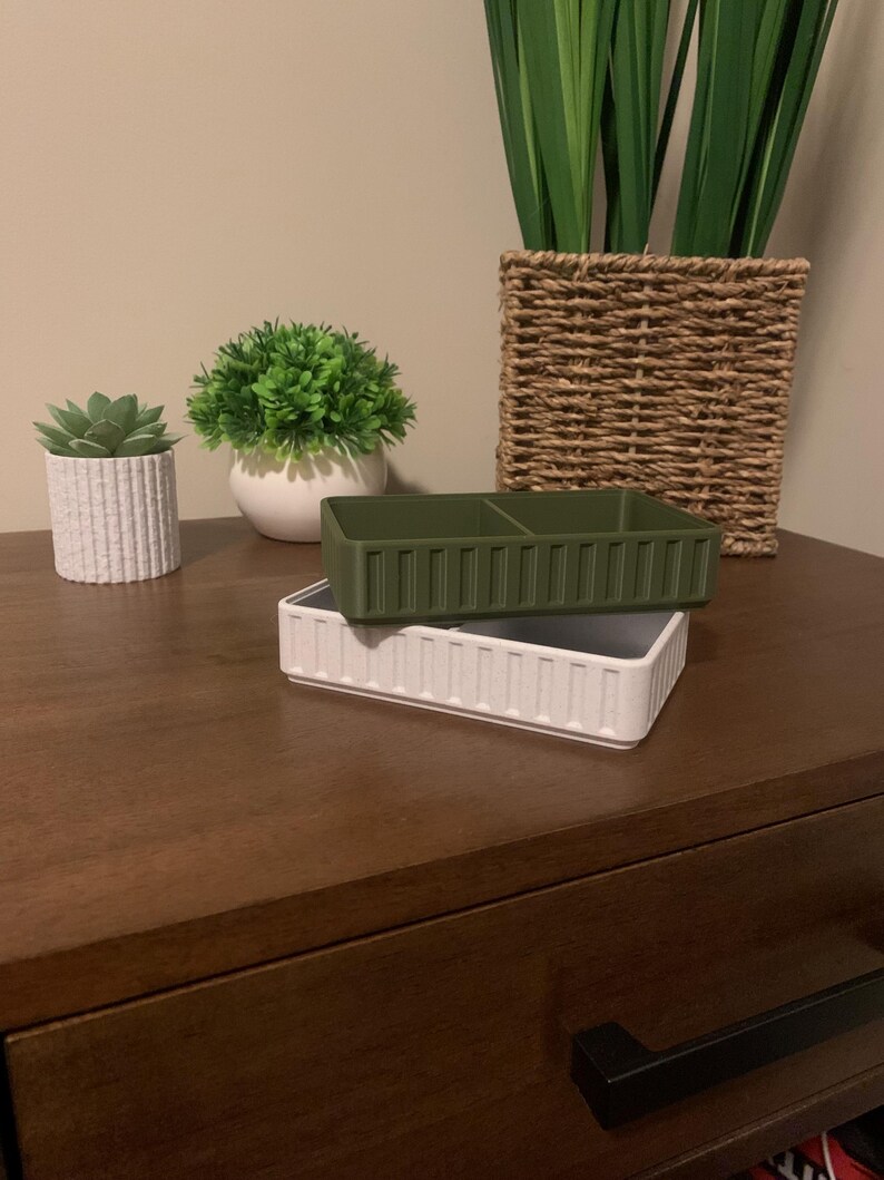 May include: Two decorative rectangular containers, one white and one olive green, are stacked on a dark wood surface. The white container has vertical ribbed detailing. The green container has two internal compartments. Artificial plants in pots are in the background.