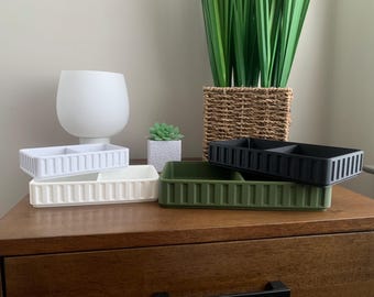 May include: A collection of rectangular storage trays in various colors: white, off-white, olive green, and black. Each tray has a divided interior and a ribbed exterior design. The trays are displayed on a wooden surface.