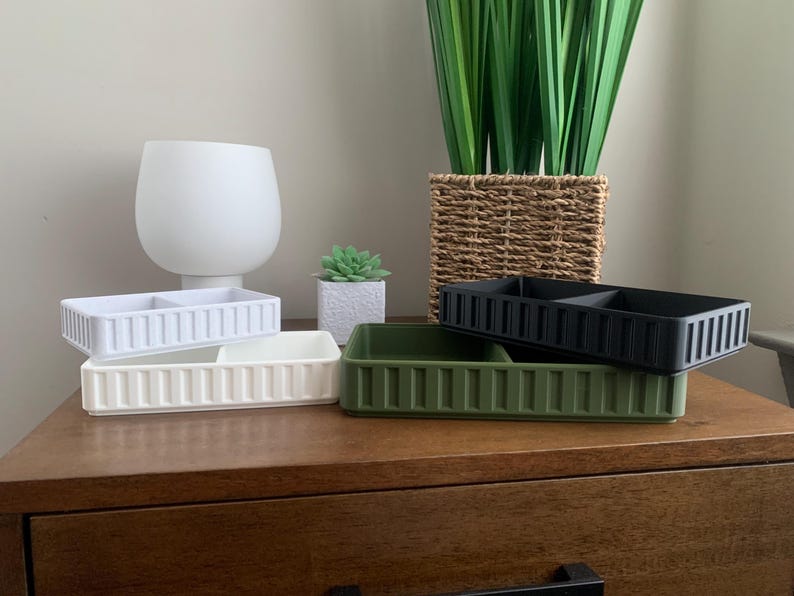 May include: Rectangular organizers in white, cream, olive green, and black, each with two compartments and vertical ribbed detailing. The organizers are stacked on a wooden surface, with a white vase, a small succulent, and a woven basket with greenery in the background.