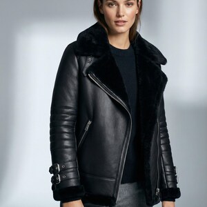 Peut inclure: Veste en cuir noire avec col et doublure en peau de mouton. La veste présente une fermeture éclair diagonale, des poignets à boucles et un style aviateur classique. Le modèle porte un pull sombre et un pantalon gris.