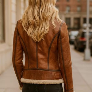 May include: A brown leather jacket with a shearling lining and trim. The jacket has a classic design with a zip closure and a fitted silhouette. The model is wearing dark jeans. The jacket is a stylish outerwear option.