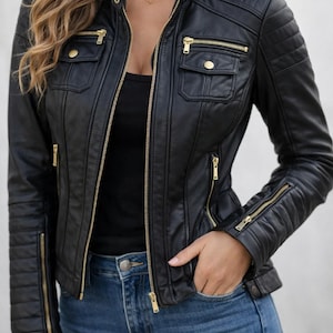 Può includere: Giacca nera in ecopelle con cerniere e dettagli dorati. La giacca presenta un design trapuntato sulle spalle e sulle maniche, due tasche sul petto e una chiusura con zip frontale. La modella indossa jeans blu.