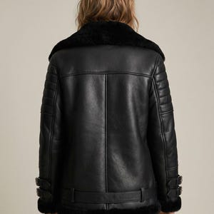 May include: A black leather jacket with a shearling collar and lining. The jacket features a belted waist, buckle details on the sleeves, and quilted shoulder panels. The back view showcases the jacket's design.