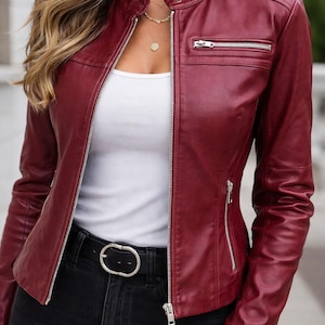 May include: A burgundy leather jacket with a stand-up collar and silver zippers. The jacket has a fitted silhouette and a zippered chest pocket. The model is wearing a white tank top and black jeans with a silver buckle belt.