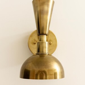 May include: A brass wall sconce with a conical top and a rounded bottom. The sconce is mounted on a circular base and has a warm, metallic finish. The design is vintage-inspired and suitable for interior lighting.