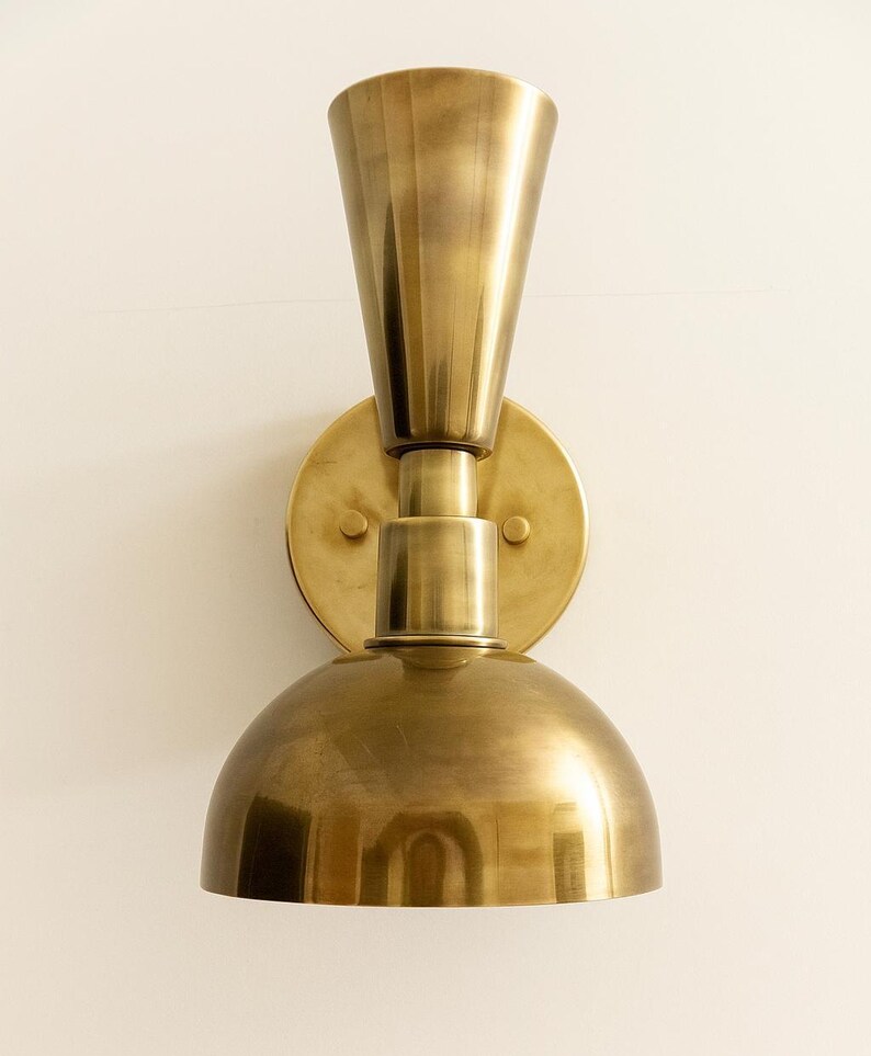 May include: A brass wall sconce with a conical top and a rounded bottom. The sconce is mounted on a circular base and has a warm, metallic finish. The design is vintage-inspired and suitable for interior lighting.