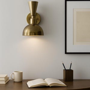 May include: A brass wall sconce with a conical top and a half-dome shade is mounted on a white wall. The sconce illuminates a wooden desk with a book, pencils, and a mug. A framed picture hangs on the wall.