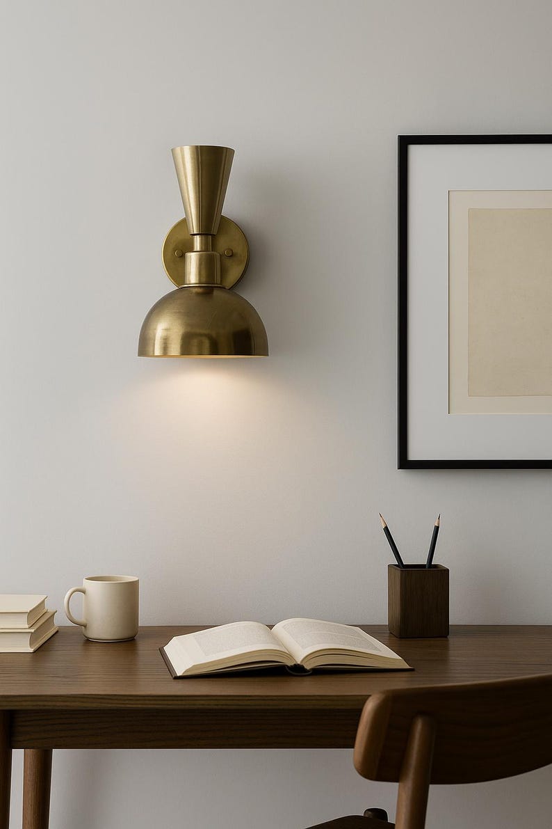 May include: A brass wall sconce with a conical top and a half-dome shade is mounted on a white wall. The sconce illuminates a wooden desk with a book, pencils, and a mug. A framed picture hangs on the wall.