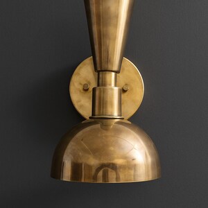 May include: A brass wall sconce with a conical top, a cylindrical midsection, and a rounded, dome-shaped bottom. The sconce is mounted on a circular backplate and has a warm, metallic finish. The fixture is designed for interior lighting.