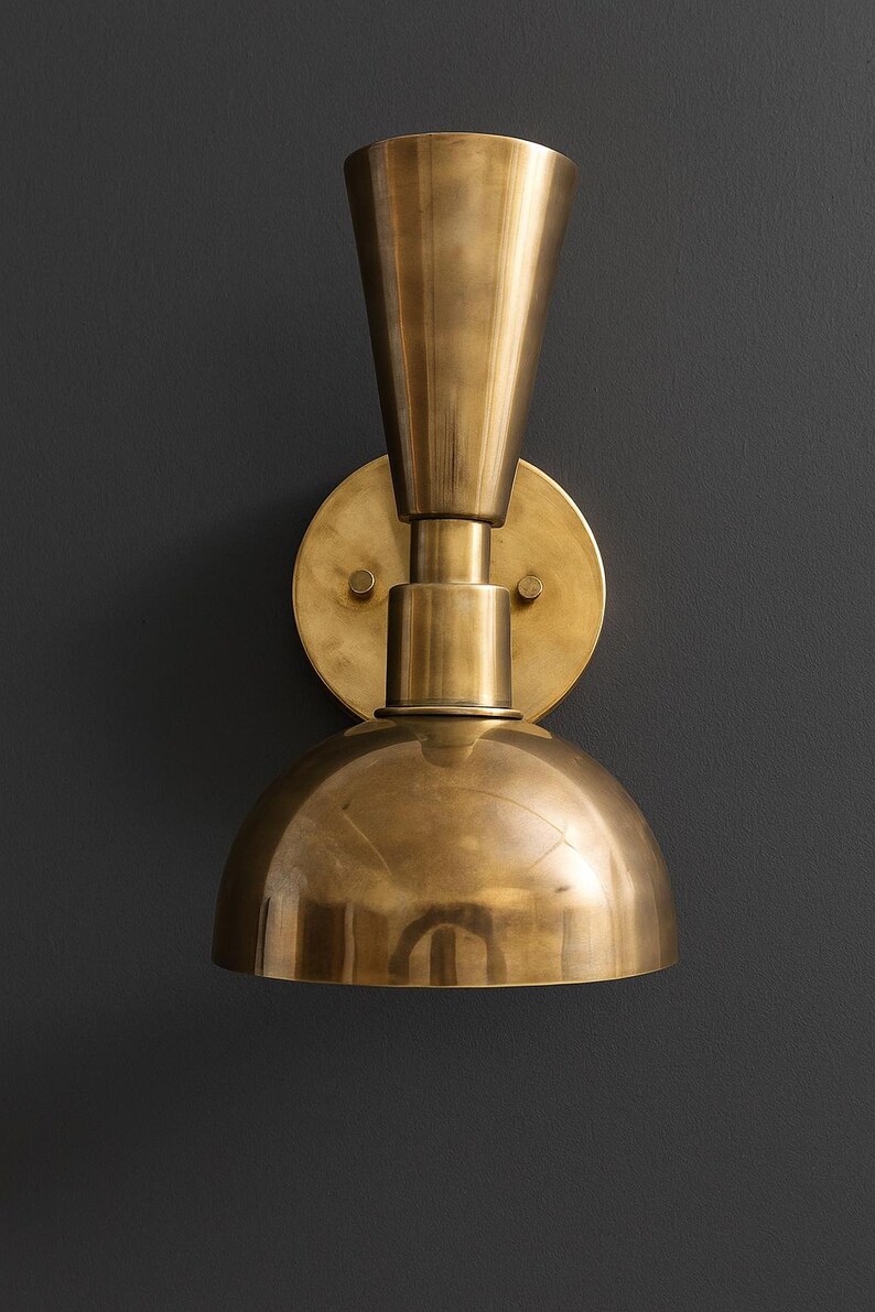 May include: A brass wall sconce with a conical top, a cylindrical midsection, and a rounded, dome-shaped bottom. The sconce is mounted on a circular backplate and has a warm, metallic finish. The fixture is designed for interior lighting.