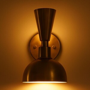 May include: A brass-toned wall sconce with a conical top and a rounded, dome-shaped bottom. The sconce is mounted on a circular backplate and emits a warm, inviting light. The design is a blend of modern and vintage styles.