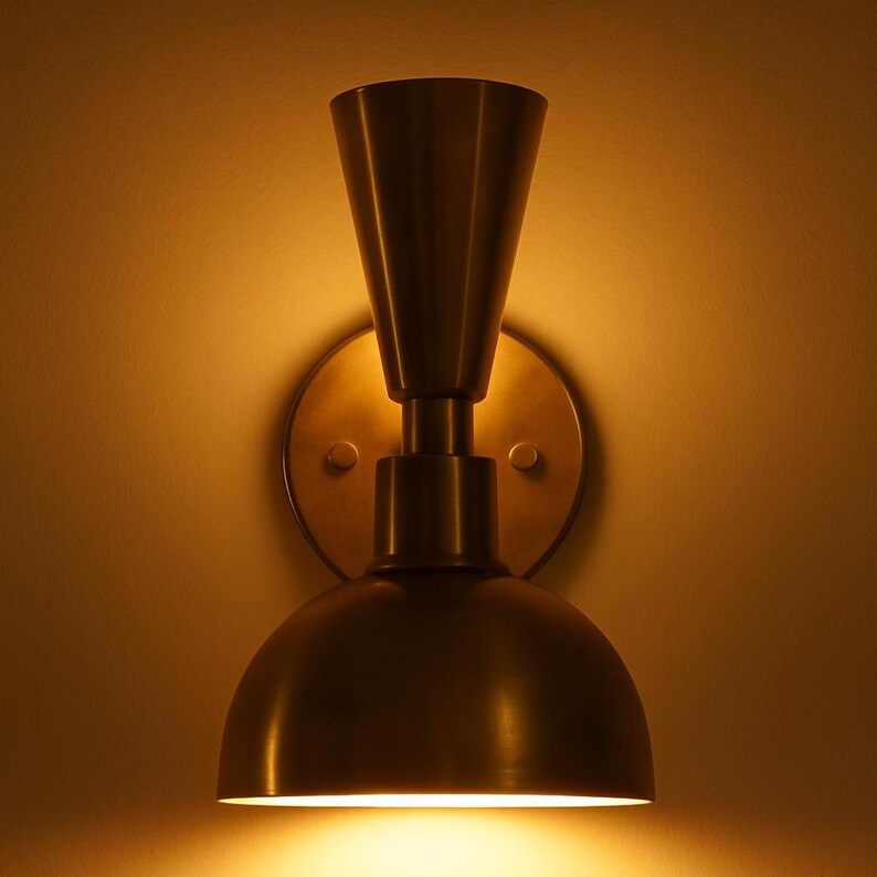 May include: A brass-toned wall sconce with a conical top and a rounded, dome-shaped bottom. The sconce is mounted on a circular backplate and emits a warm, inviting light. The design is a blend of modern and vintage styles.