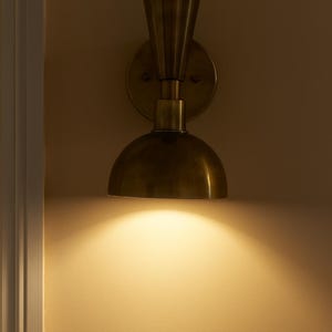 May include: A brass wall sconce with a half-dome shade and a fluted top. The sconce is mounted on a circular backplate and casts a warm, downward light. The fixture is a vintage-inspired design.