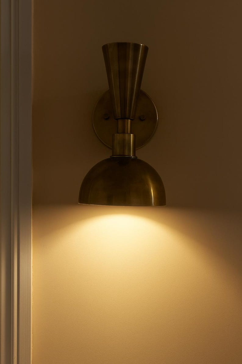 May include: A brass wall sconce with a half-dome shade and a fluted top. The sconce is mounted on a circular backplate and casts a warm, downward light. The fixture is a vintage-inspired design.