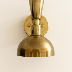 May include: A brass wall sconce with a conical top and a rounded, dome-shaped bottom. The sconce is mounted on a circular backplate with two small screws. The fixture has a warm, metallic finish.