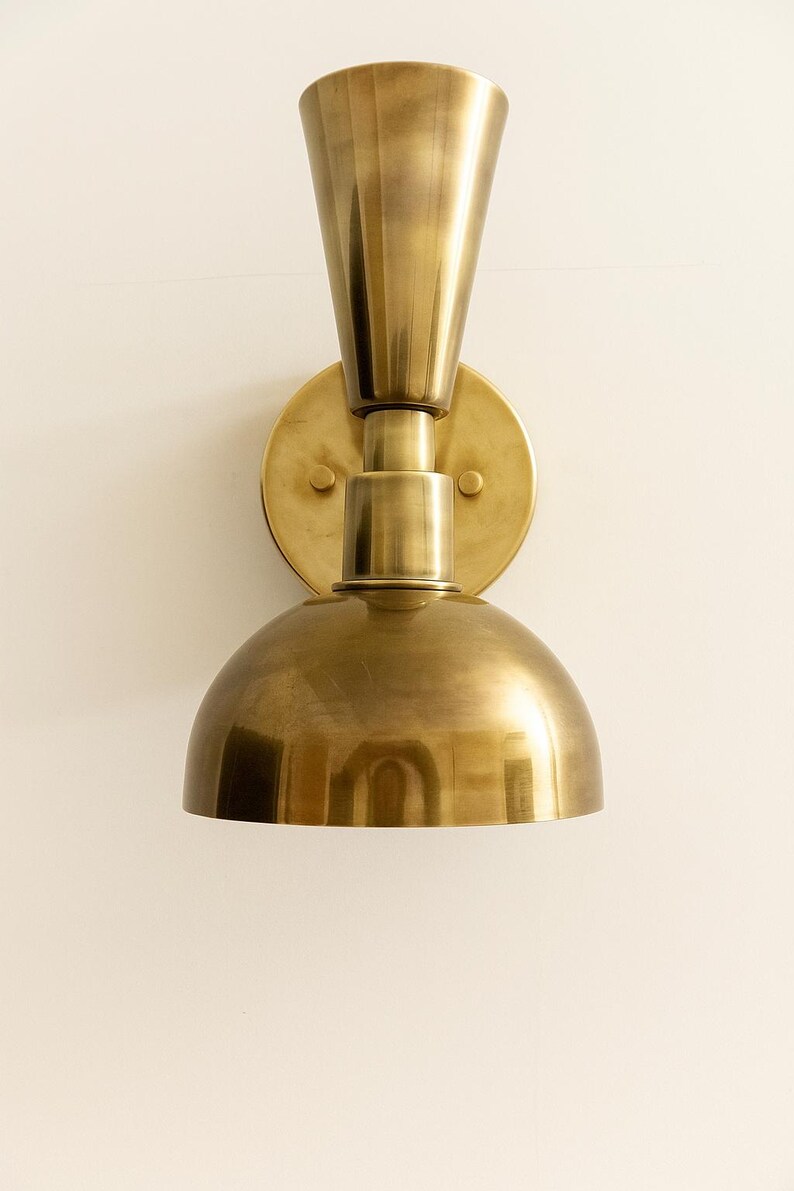 May include: A brass wall sconce with a conical top and a rounded, dome-shaped bottom. The sconce is mounted on a circular backplate with two small screws. The fixture has a warm, metallic finish.