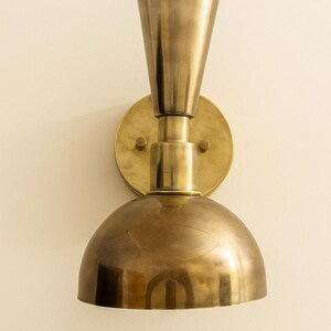 May include: A brass wall sconce with a conical top and a rounded, dome-shaped bottom. The sconce is mounted on a circular backplate and has a warm, metallic finish. The design is a vintage style.