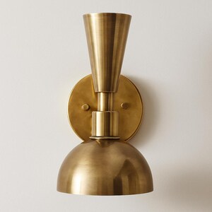 May include: A brass wall sconce with a conical top, a circular backplate, and a rounded bottom. The sconce has a vintage aesthetic and is mounted on a white wall. The fixture is made of metal and has a warm, golden hue.