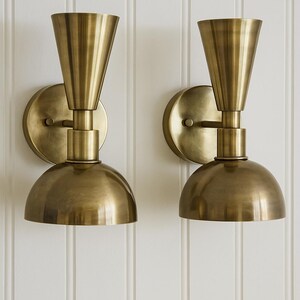 May include: Two brass wall sconces with a cone-shaped top and a rounded bottom. The sconces are mounted on a circular base and have a warm, metallic finish. The background is a white, vertical-paneled wall.