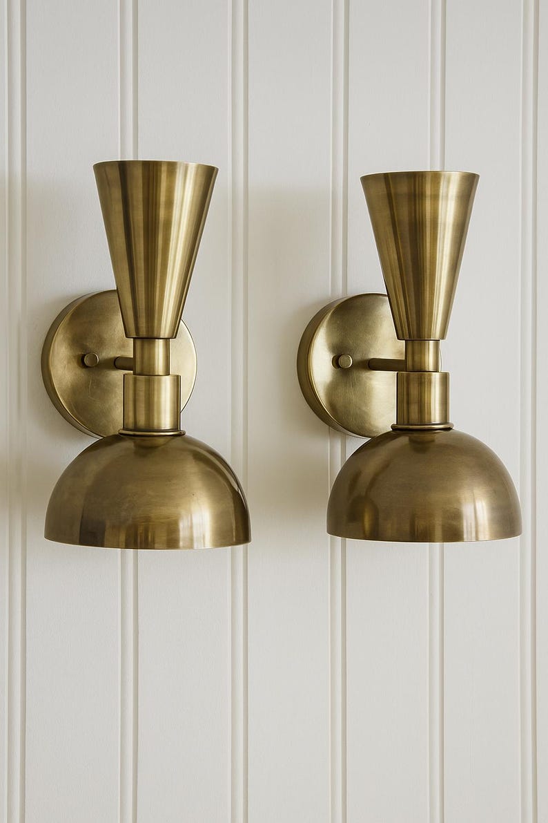 May include: Two brass wall sconces with a cone-shaped top and a rounded bottom. The sconces are mounted on a circular base and have a warm, metallic finish. The background is a white, vertical-paneled wall.